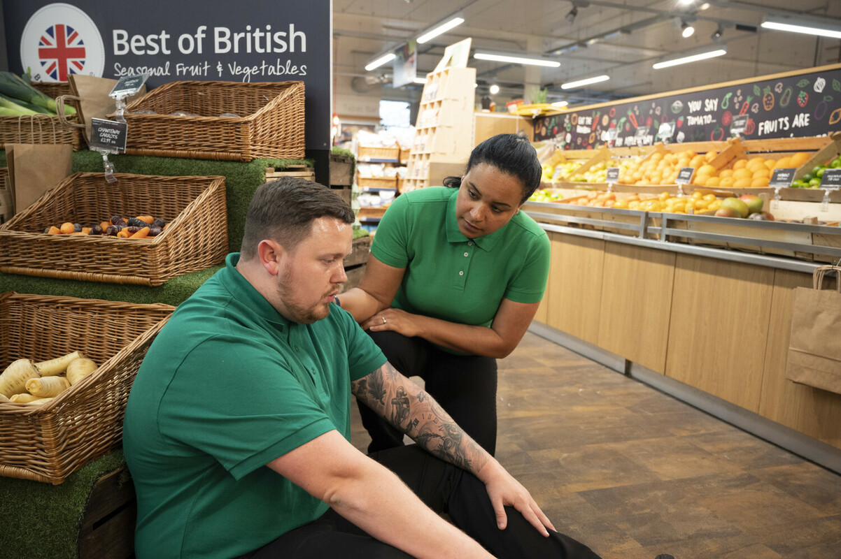 A first aider in a supermarket is helping a colleague, who is sat on the floor doing deep breathing.